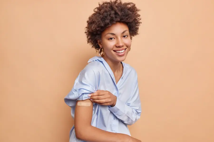 Pretty pleased Afro American woman in blue shirt raises sleeve shows vaccinated arm wears adhesive plaster smiles pleasantly isolated over beige wall