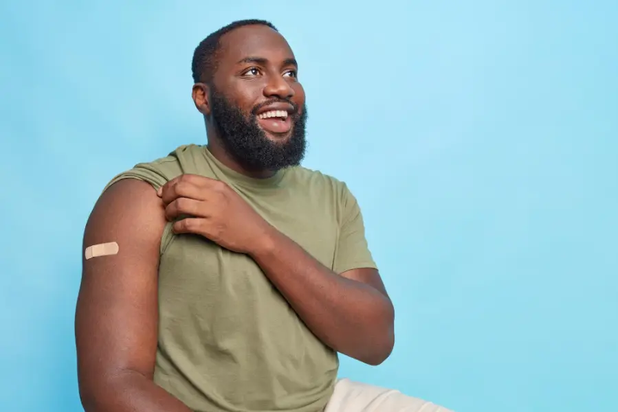 Vaccination campaign against covid 19. Cheerful dark skinned man shows arm with adhesive plaster after being vaccinated receives vaccine poses against blue copy space area for your text