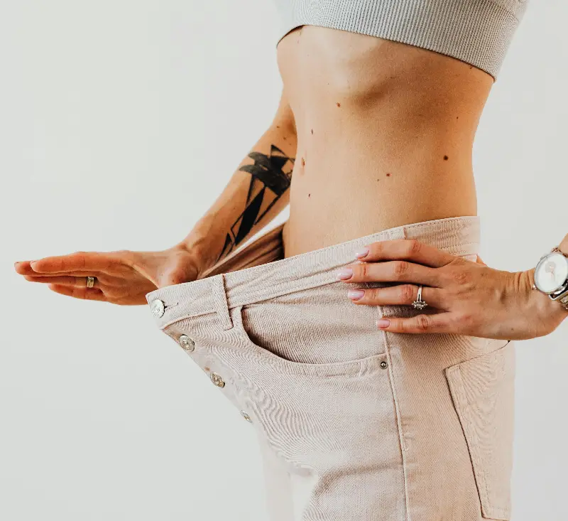 Close-up of a woman in oversized pants showcasing weight loss progress. Studio shot with neutral background.