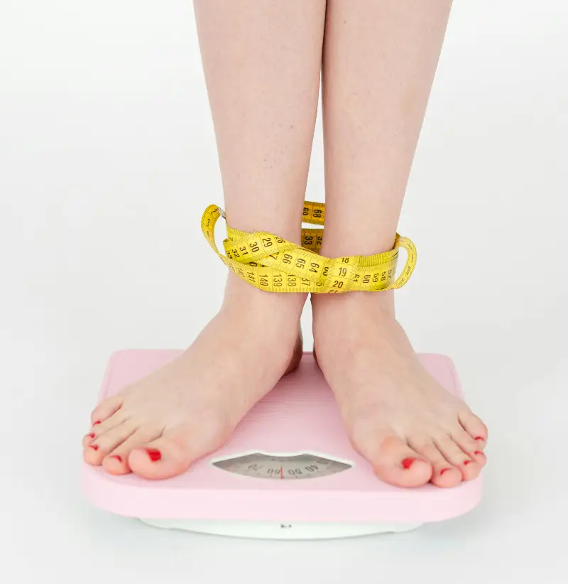 High angle of crop anonymous female with centimeter on legs checking weight on scales on white background